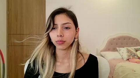 Media: Video of a young Latina woman with long, straight, blonde-highlighted hair, wearing a black top and gold earrings, in a beige bedroom with a white bed and floral pillows.