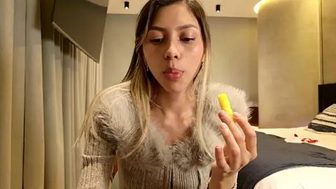 Media: Video of a Latina woman with long, straight, blonde hair, wearing a fur-trimmed, beige sweater, blowing a yellow bubble gum, in a modern, warmly lit bedroom with beige curtains and a bed.