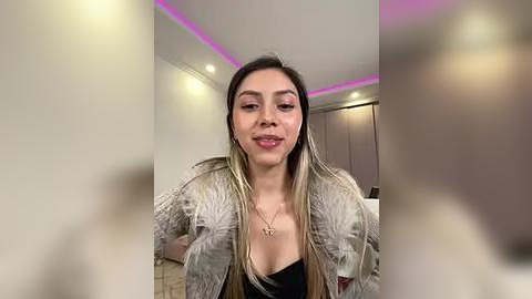 Media: Video of a smiling Latina woman with long, straight, blonde hair, wearing a black top and a silver feathered jacket, standing in a modern, well-lit room with white walls and pink accent lighting.