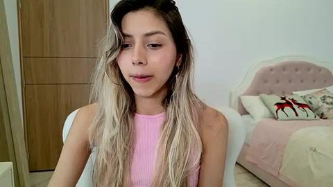 Media: Video of a young woman with long, wavy blonde hair, wearing a sleeveless pink top, sitting on a bed with a pink headboard and decorative pillows in a minimalist bedroom.