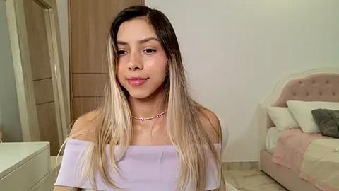 Media: Video of a young woman with long, straight, ombre hair, wearing a light purple off-shoulder top, standing in a simple, modern bedroom with a beige bed and wooden door.