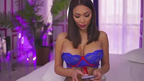 Media: Video of a Latina woman with straight black hair, wearing a blue lace bra, holding a smartphone, in a modern, dimly lit bedroom with purple accents and potted plants.