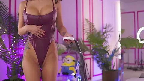 Media: Video of a woman with fair skin and long brown hair, wearing a tight, metallic maroon bodysuit with plunging neckline, holding a white smartphone. Background features tropical plants, a Minion statue, and a studio light.