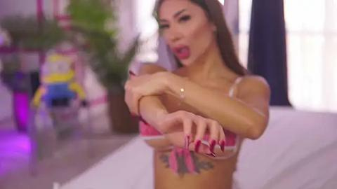 Media: Video of an Asian woman with long brown hair, wearing a pink bra, stretching, with a tattoo on her left ribcage, in a brightly lit bedroom with green potted plants and colorful toys in the background.