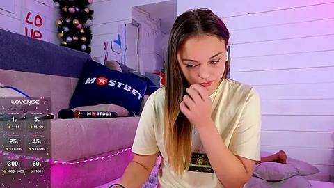 Media: A young, slender woman with long brown hair, wearing a white t-shirt, sits pensively on a bed in a cozy room. The room is decorated with a Christmas tree and a \"LOVE\" sign.