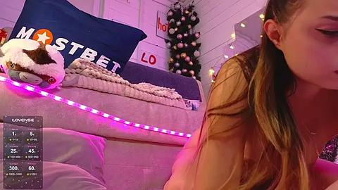 Media: A video of a young woman with long brown hair, wearing a brown top, sitting on a bed with a Christmas tree in the background, illuminated by purple string lights.