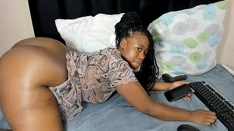 Media: Video of a Black woman with braided hair, wearing a patterned top, lying on a bed with a floral pillow, using a laptop, revealing her buttocks.