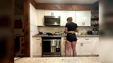 Media: Video of a woman with a ponytail, wearing a black crop top and shorts, cooking in a modern, white kitchen with stainless steel appliances.