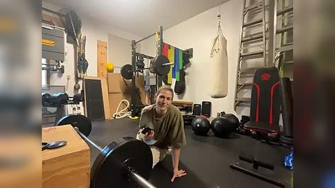 Media: A video of a smiling woman in workout gear, sitting on a gym floor surrounded by various exercise equipment, including weights, a punching bag, and a wooden crate.