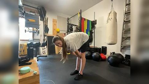 Media: Video of a young woman with long brown hair in a gym, stretching, wearing a gray shirt and black shorts. Background includes weights, a punching bag, and various workout equipment.