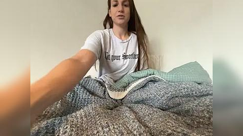 Media: Video of a young woman with long brown hair, wearing a white T-shirt with black text, sitting on a bed with a textured gray blanket.