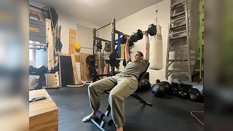 Media: Video of a muscular man in a gym, lifting a barbell with a white towel, wearing green workout clothes. Background includes various weights, punching bags, and wooden structures.