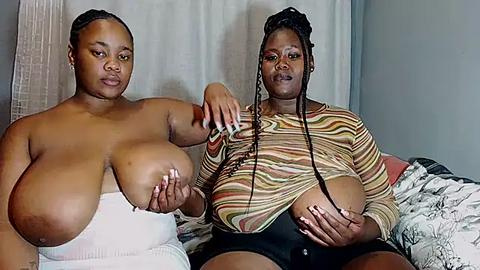 Media: Video of two large-breasted Black women, one topless, the other wearing a striped top, sitting on a bed with patterned sheets.