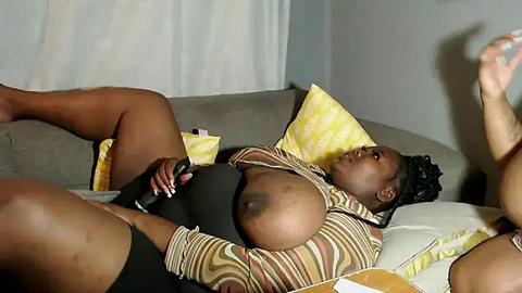 Media: Video of a Black woman with dark skin, large breasts, and curly hair, lying on a gray couch, wearing a striped crop top, black shorts, and a yellow pillow.
