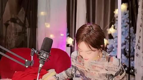 Media: Video of a woman with brown hair, wearing a newspaper-print top, singing into a microphone on a red couch, surrounded by festive decorations, including a Christmas tree.