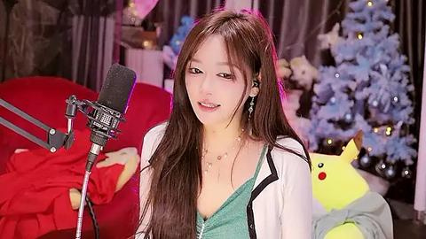 Media: Video of a young Asian woman with long brown hair, smiling, wearing a green top, seated in front of a microphone, surrounded by festive decor, including a Pikachu plush and a Christmas tree.