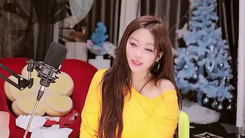 Media: Video of a smiling young Asian woman with long brown hair, wearing a yellow off-shoulder top, sitting in front of a microphone on a red couch. Christmas tree and fluffy toys in the background.