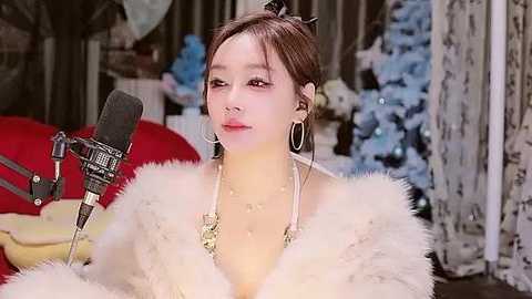 Media: Video of a young Asian woman with light skin and brown hair, wearing white fur, a gold necklace, and hoop earrings, singing into a microphone in a cozy, softly lit room.