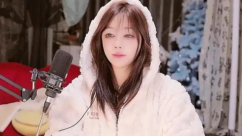 Media: Video of an Asian woman with straight brown hair in a white fluffy hoodie, holding a microphone, standing indoors with a cluttered background.