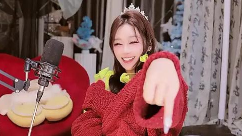 Media: A video of an Asian woman with long brown hair and a tiara, smiling widely, wearing a red sweater, pointing towards a microphone on a red cushion. Background features a floral curtain and various household items.