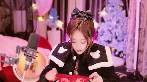 Media: Video of an East Asian woman with brown hair, dressed in a black, fur-trimmed jacket, reading a book. Background features a Christmas tree, pink and white lights, and a microphone on a stand.