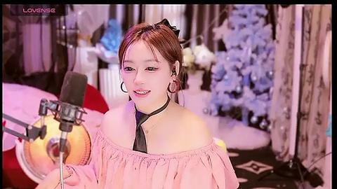 Media: Video of a smiling Asian woman with light skin, brown hair, wearing a pink off-shoulder top and black tie, microphone in hand, recording in a cozy, softly lit room with a white Christmas tree and festive decor.