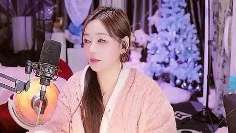 Media: Video of a young East Asian woman with long brown hair, wearing a beige cardigan, singing into a microphone, set against a winter-themed backdrop with snowflakes and a Christmas tree.