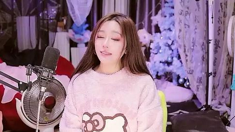 Media: Video of a young Asian woman with long brown hair, wearing a white sweater with a cartoon cat design, singing into a microphone in a cozy, softly lit room with a decorated Christmas tree in the background.