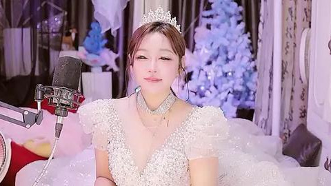 Media: Video of a young East Asian woman with light skin, wearing a white, beaded, puffed-sleeve dress, a silver tiara, and a choker, singing into a microphone, surrounded by festive, white and blue decorations.