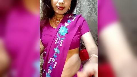Media: Video of an Indian woman in a vibrant magenta sari with intricate blue and white floral embroidery, wearing gold jewelry, standing in a tiled room with a blurred background.