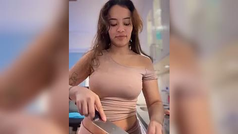 Media: Video of a young woman with light brown skin and long wavy hair, wearing a tight, off-shoulder beige top, cutting a piece of paper with scissors in a modern kitchen.