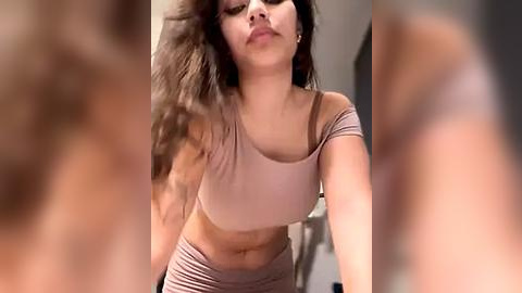 Media: Video of a young woman with long, wavy brown hair, wearing a beige off-shoulder crop top and matching high-waisted pants. Background is blurred, with an indistinct indoor setting.