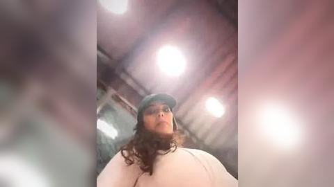 Media: Video of a woman with curly hair and a green cap, standing indoors under bright lights, wearing a beige sweater, blurred background.