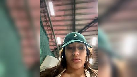 Media: Video of a young woman with medium-brown skin and long brown hair, wearing a green baseball cap, green shirt, and brown jacket, standing in an industrial warehouse with exposed metal beams and green walls.