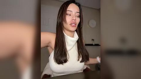 Media: Video of a young woman with straight, long dark hair, wearing a white sleeveless top, leaning over a table in a dimly lit room with beige walls and a white clock.