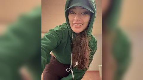 Media: Video of a young woman with long brown hair, wearing a green hooded jacket, smiling, blurred background, indoors.