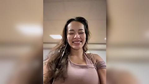 Media: Video of a joyful young woman with long, wavy dark hair, wearing a light pink tank top, smiling with eyes closed, standing in a modern, softly lit indoor setting with blurred background.