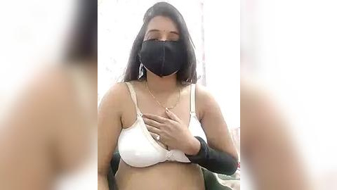 Media: Video of a young woman with medium skin tone, wearing a white bra and black face mask, holding her chest.