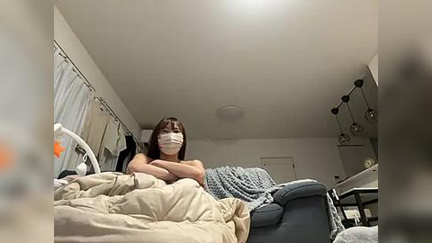 Media: Video of a young woman with straight, shoulder-length brown hair, wearing a white face mask and a black top, sitting on a beige bed in a dimly lit, cluttered bedroom.