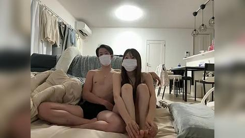 Media: Video of a young East Asian couple wearing face masks, sitting closely on a beige sofa in a modest, dimly lit living room.