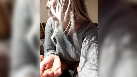 Media: A video of a woman with long, blonde hair, wearing a grey sweater, seated on a couch. Her hands are extended forward, fingers slightly curled. The background is blurred, creating a soft focus effect.