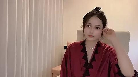 Media: Video of a young East Asian woman with fair skin and dark hair tied back, wearing a maroon silk robe with black frills, seated against a beige wall with vertical blinds.