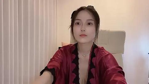 Media: A video of an Asian woman with fair skin, dark hair in a bun, wearing a red satin robe with black lace trim, seated on a white chair against a beige wall with vertical blinds.