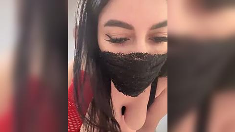 Media: Video of a woman with long black hair and dark, arched eyebrows, wearing a black lace face mask and a red top, with a blurred background.