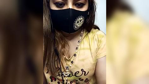 Media: Video of a young woman with light brown skin, wearing a black mask, gold chain necklace, and yellow t-shirt with \"coffee\" printed in brown. Background is blurred.