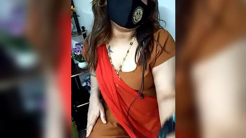 Media: Video of a woman with wavy brown hair, wearing a red sari, black mask, and a gold chain, sitting indoors.