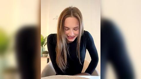 Media: Video of a young Caucasian woman with long, straight, light brown hair, wearing a black long-sleeve top, smiling while leaning over a table in a modern, light-colored room.