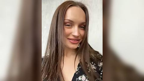 Media: Video of a young woman with long, straight brown hair, fair skin, and a subtle smile. She wears a black floral dress with white flowers. The background is a plain, light-colored wall.