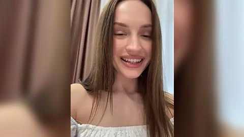 Media: Video of a young Caucasian woman with long, straight brown hair, wearing a white off-shoulder blouse, smiling joyfully. Background shows blurred, muted colors.