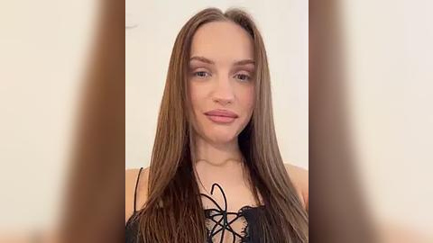 Media: Video of a young Caucasian woman with long, straight brown hair and light skin, wearing a black lace-up top, smiling subtly. The background is a plain white wall, slightly blurred.